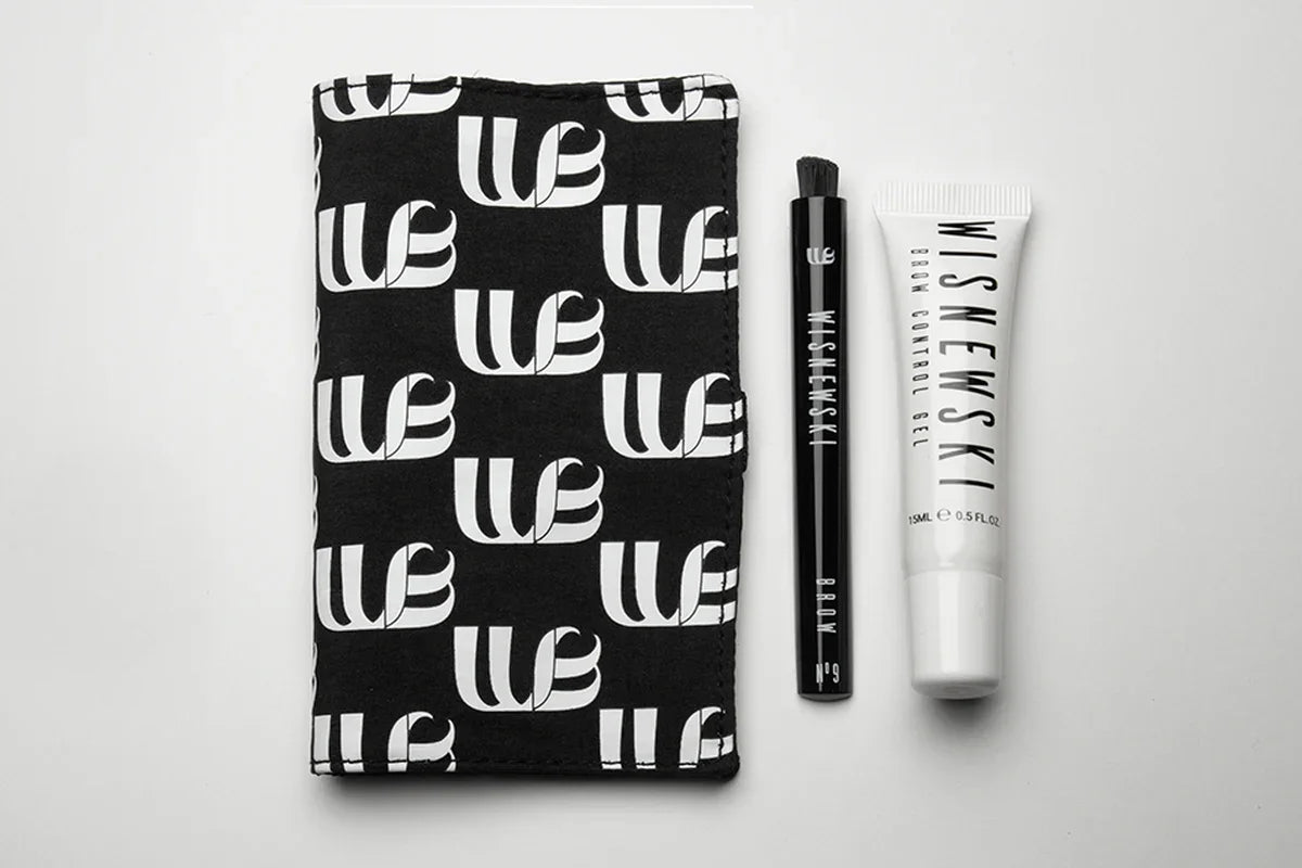 Outside view of the Wisnewski Magnetic Pouch. The pouch is black with a white Wisnewski logo printed in a tiled pattern. There is also one of the #9 Eyebrow dry-brushes and a white tube of the Extra Strong Control Gel to the right of the magnetic pouch.